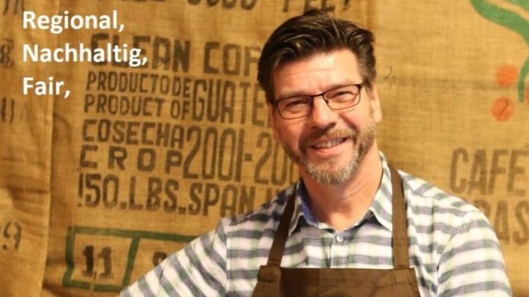 Person in brauner Schürze mit Aufschrift „Die Mainzer Kaffeemanufaktur“ hält Schaufel mit grünen Kaffeebohnen vor Kaffeesäcken.