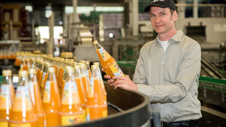 Eine Person hält eine Flasche mit orangefarbener Flüssigkeit vor einer Produktionslinie mit weiteren Flaschen.