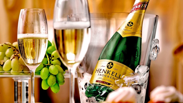 Eine Henkell-Sektflasche in einem Eiskübel, zwei gefüllte Sektgläser, grüne Weintrauben und Dessert auf weißem Tisch.