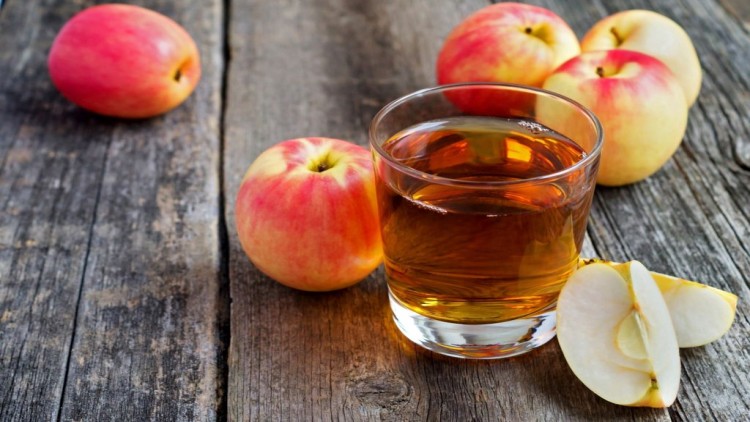 Ein Glas klarer Apfelsaft steht auf einer Holzoberfläche, umgeben von roten Äpfeln und zwei Apfelscheiben.