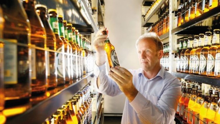 Person in einem Hemd hält eine Bierflasche in der Hand und steht zwischen Regalen mit weiteren Bierflaschen.