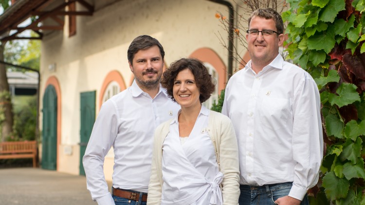 Drei Personen in weißer Kleidung und Jeans stehen vor einem Gebäude mit grünen Fensterläden und Weinranken.