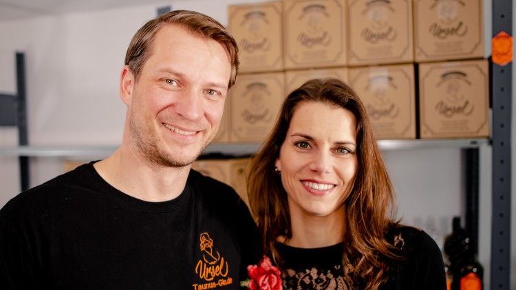 Ein Mann trägt ein schwarzes T-Shirt mit orangefarbenem Logo und Schriftzug „Ursel Taunus-Gin.de“, im Hintergrund stehen Kartons.