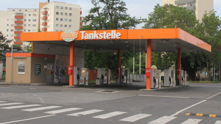 Eine GLOBUS Tankstelle mit orangefarbenem Dach, mehreren Zapfsäulen und einem Stoppschild im Vordergrund.
