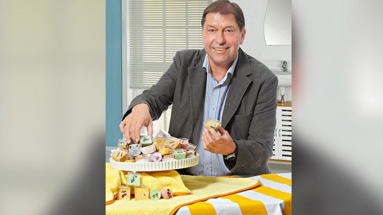 Person in Anzug legt eine Mini-Torte auf eine weiße Etagere mit verschiedenen kleinen Kuchen, gelbes Tuch im Hintergrund.