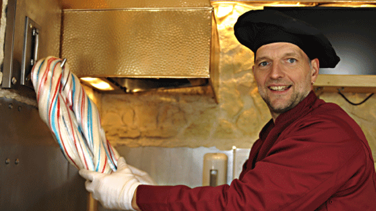 Person trägt Handschuhe und hält eine mehrfarbige Zuckerstange in einer Backstube vor einem goldfarbenen Abluftsystem.
