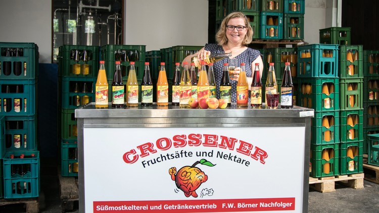 Mehrere Flaschen Crossener Fruchtsäfte und Nektare auf einem Verkaufstresen mit Werbetafel, daneben grüne Getränkekisten.