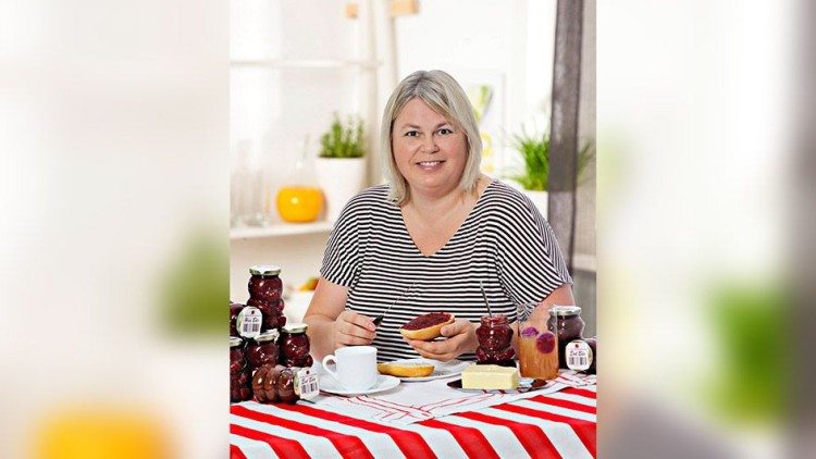 Person streicht Marmelade aus einem Glas auf ein Brötchen, umgeben von weiteren Marmeladengläsern, Tee und Butter auf einem Tisch.