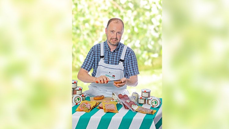 Person in weißer Schürze an einem Tisch mit verpackten Wurstspezialitäten, Aufstrichen und Gläsern vor unscharfem Hintergrund.