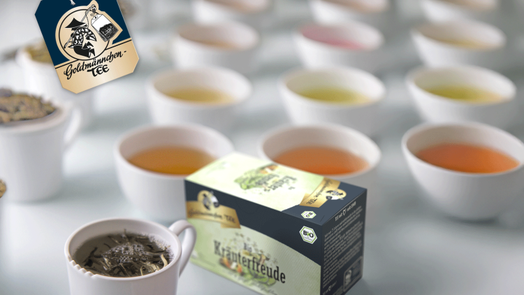 Verschiedene Teetassen mit buntem Tee, eine Teebeutel-Aufschrift „Goldmännchen Tee“ und schwarze Kräuterfreude-Teepackung.