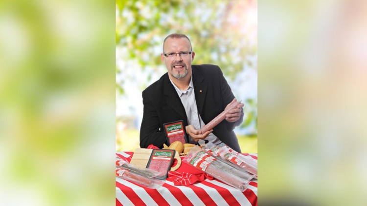 Person in dunklem Sakko zeigt eine leicht verpackte Wurst über einem Tisch mit gestreifter Tischdecke und weiteren Würsten.