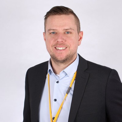 Person in schwarzem Sakko, hellblauem Hemd und mit gelbem Schlüsselband vor neutralem Hintergrund.