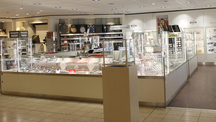 Schmuck- und Uhrenverkaufsbereich mit beleuchteten Glasvitrinen, Hinweistafeln und Schildern wie „Uhren-Service“ und „sale“.