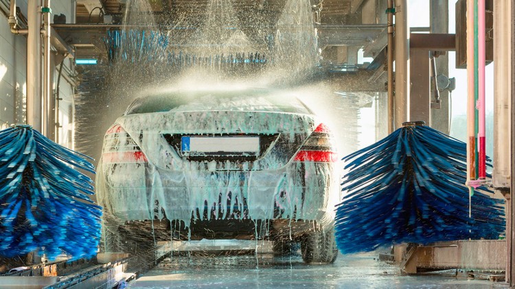 Ein Auto wird in einer automatisierten Waschanlage mit Wasser und Schaum gereinigt, blaue Bürsten sind seitlich sichtbar.