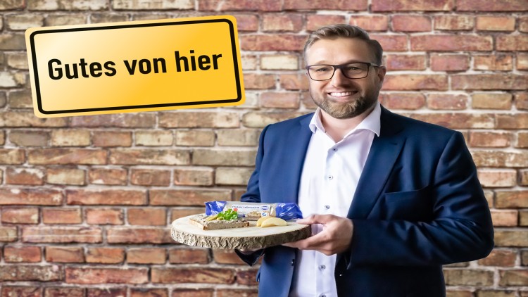 Person in blauem Anzug hält Holzplatte mit Leberwurst, Brot, Petersilie und Apfelscheiben, Schild mit „Gutes von hier“ darüber.