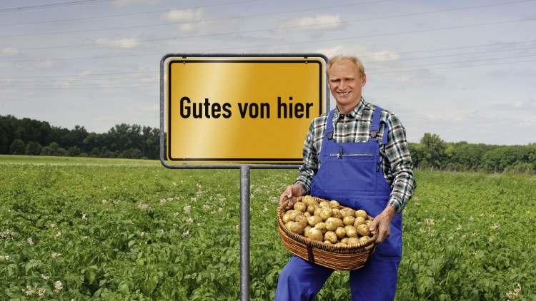 Ein Mensch mit blauer Latzhose hält einen Korb mit Kartoffeln vor einem Feld, daneben ein Schild mit der Aufschrift Gutes von hier.