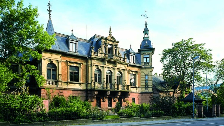 Historisches Gebäude mit verzierten Fassaden, spitzem Turm, großer Eingangstür und umliegenden Bäumen an einer Straße.