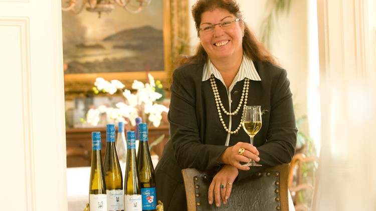 Eine Person in dunklem Blazer hält ein Glas Weißwein, daneben stehen mehrere Weinflaschen mit blauem Verschluss auf einem Tisch.