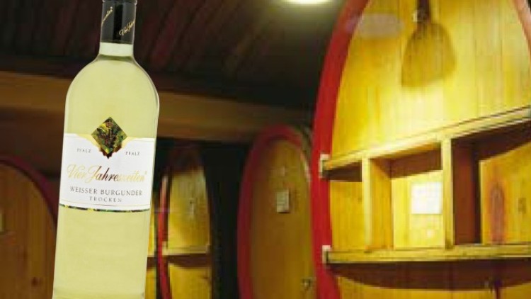 Eine Flasche Weißwein mit der Aufschrift „Villa Jahnweiler, Weisser Burgunder, trocken“ steht vor Holzfässern in einem Weinkeller.