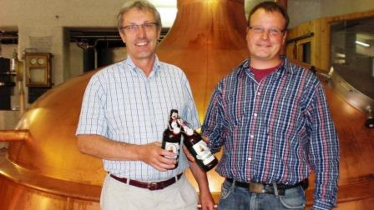 Zwei Personen in karierter Kleidung halten Bierflaschen vor einem großen, kupferfarbenen Braukessel in einer Brauerei.