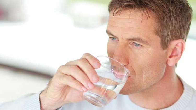 Person in weißem Pullover hält ein Glas Wasser in der rechten Hand und trinkt daraus, Hintergrund unscharf.