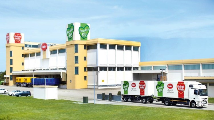 Gelbes Fabrikgebäude mit Schildern „Paradiso Bio“ und „Frohsnack Rubin“, davor Lkw mit passenden Aufschriften auf Anhängern.