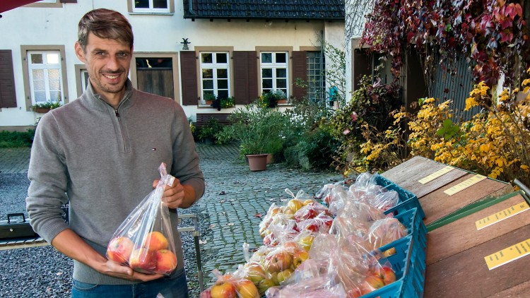 Person hält eine Plastiktüte mit Äpfeln in der Hand, daneben blaue Kisten mit weiteren abgepackten Äpfeln auf einem Tisch.