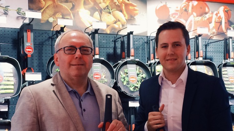 Zwei Personen halten Pfannen der Marke WOLL mit Speisen, im Hintergrund eine Produktwand mit weiteren Pfannen.