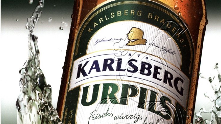 Eine braune Bierflasche mit Karlsberg-UrPils-Etikett wird von spritzendem, klaren Wasser umgeben.