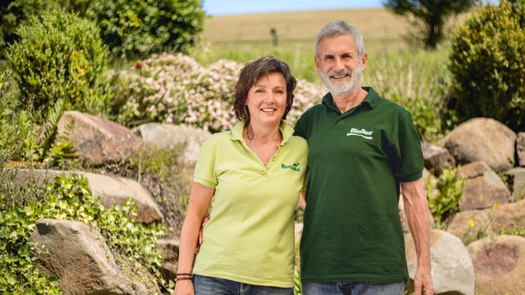 Zwei Personen in grünen Polohemden stehen vor einer Gartenlandschaft mit Steinen, Blumen und Sträuchern.