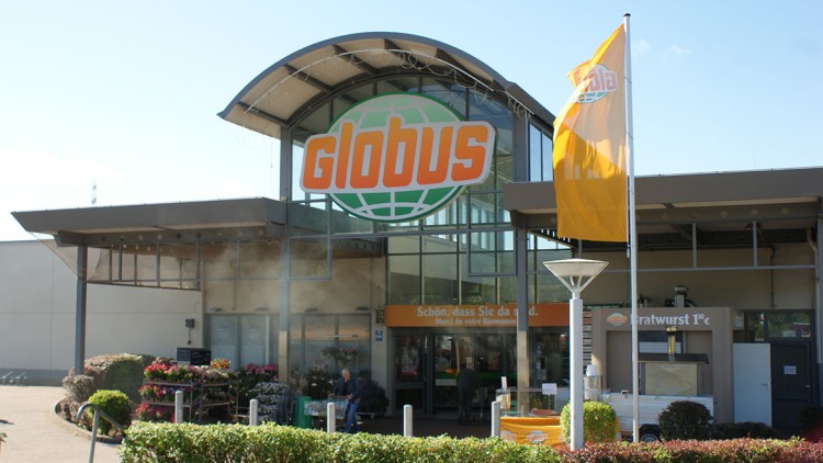 Eingang eines GLOBUS Marktes mit großem Logo, Blumenauslage, orangem Willkommensbanner und gelber Fahne im Vordergrund.