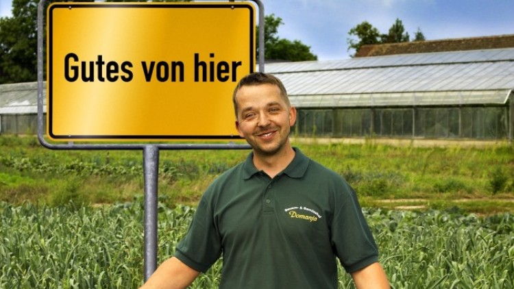 Ein Mann in grüner Arbeitskleidung präsentiert frisches Gemüse vor einem Schild mit der Aufschrift Gutes von hier.