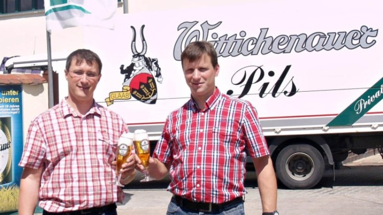 Zwei Personen in karierten Hemden stoßen mit Biergläsern an, im Hintergrund ein LKW mit „Wittichenauer Pils“-Schriftzug.