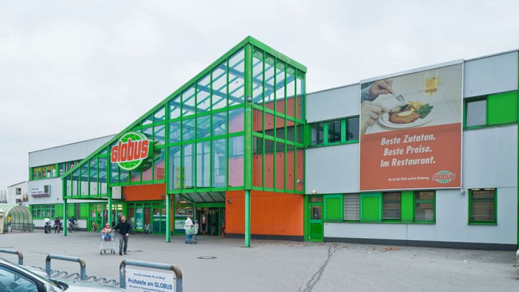 Eingang eines GLOBUS-Marktes mit markantem Glasdach, geparkten Autos und Werbeplakat mit dem Text „Beste Zutaten, Beste Preise, Im Restaurant“.