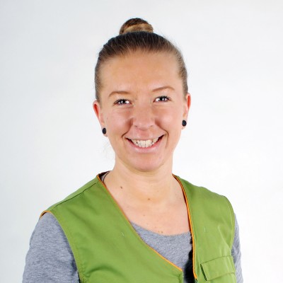 Person trägt eine grüne Weste mit orangener Kante über einem grauen Oberteil, Hintergrund ist einfarbig weiß.