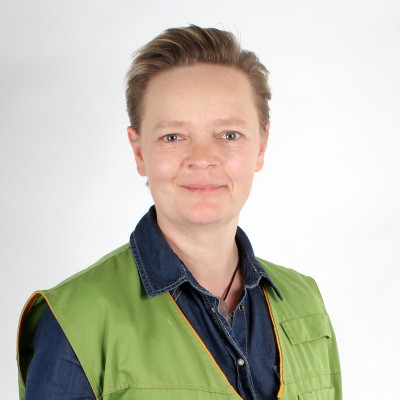 Person in grüner Weste mit orangener Umrandung und dunklem Jeanshemd vor grauem Hintergrund.