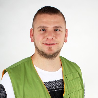 Person trägt eine grüne Weste mit orangen Nähten und ein weißes Shirt mit schwarzem Muster vor einem weißen Hintergrund.