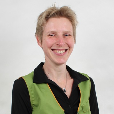Person trägt schwarze Bluse, grüne Weste mit orangen Nähten und eine Halskette mit rechteckigem Anhänger.