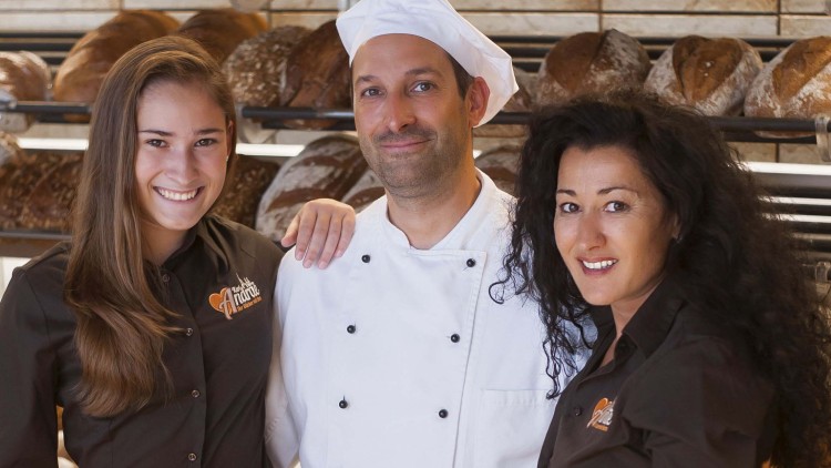Drei Personen in Bäckeruniformen stehen vor Regalen mit verschiedenen Brotlaiben.