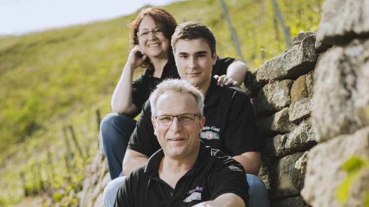 Drei Personen in schwarzen Polo-Shirts mit Aufschrift „Wein-Gut“ sitzen vor einer Steinmauer in einer Weinberg-Landschaft.
