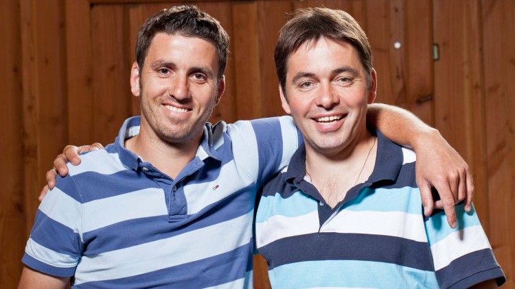 Zwei Personen in gestreiften Poloshirts stehen vor einer Holzwand, eine legt den Arm um die Schulter der anderen.
