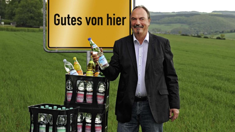 Ein Mann in Anzug steht neben Getränkekisten mit Wasserflaschen, im Hintergrund ein Schild mit der Aufschrift Gutes von hier.