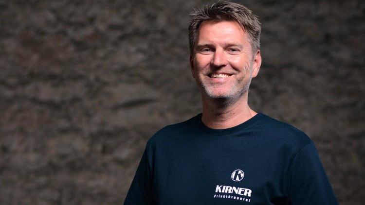 Person in dunkelblauem T-Shirt mit weißem Logo und Schriftzug „KIRNER Privatbrauerei“ vor unscharfer Steinwand.