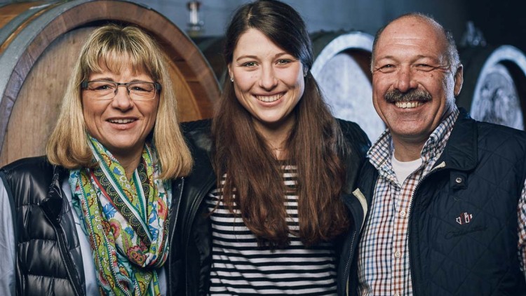 Drei Personen stehen vor großen Holzfässern, tragen Freizeitkleidung mit Westen und Schals, unscharfer Hintergrund innen.