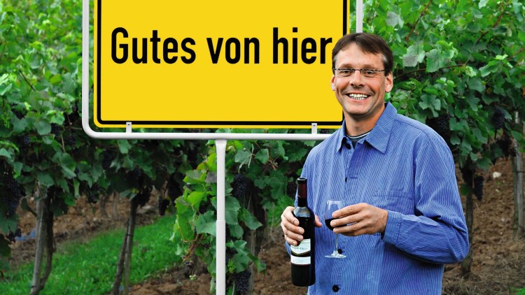Person vor Weinreben hält eine Weinflasche und ein Weinglas, gelbes Schild im Hintergrund mit der Aufschrift Gutes von hier.