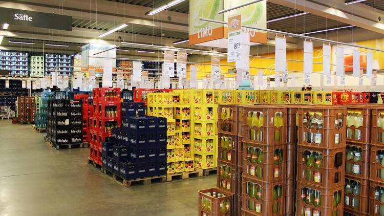 Getränkekisten in verschiedenen Farben stehen auf Paletten, darüber Schilder mit Preisen, Bereich mit „Säfte“ und „Limo“ gekennzeichnet.