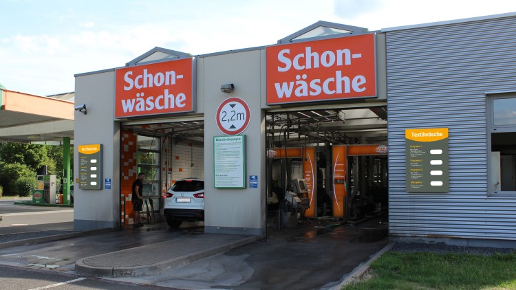 Zwei geöffnete Tore einer Autowaschanlage mit der Aufschrift „Schonwäsche“, ein Auto fährt in die linke Spur.