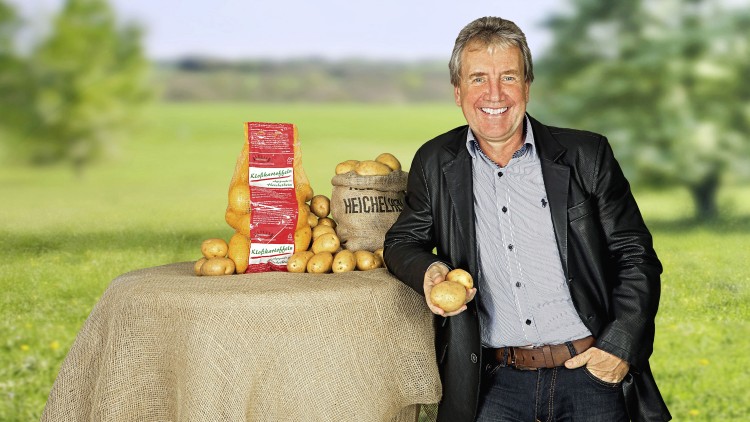 Ein Tisch mit Kartoffeln, einem Kartoffelsack mit Schriftzug „Heichelheimer“ und einer roten Kartoffelverpackung steht im Freien.
