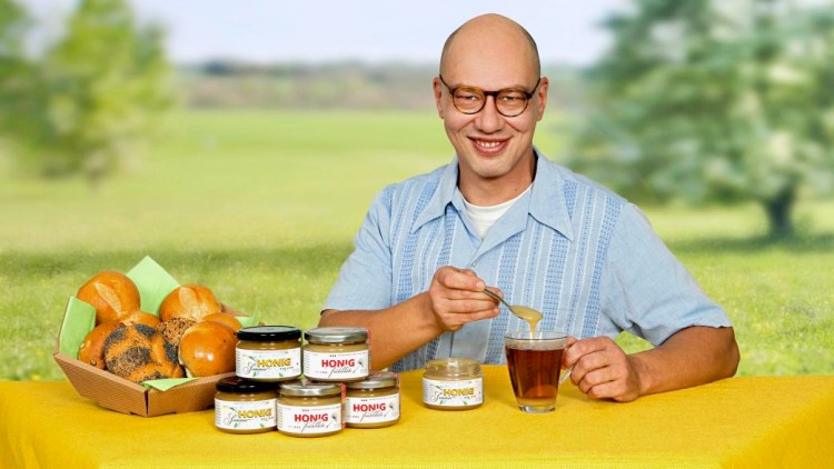 Auf gelber Tischdecke stehen Brötchen, verschiedene Honiggläser und ein Glas Tee, in das Honig mit einem Löffel gegeben wird.