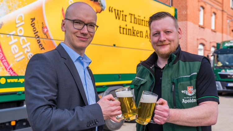 Zwei Personen stoßen mit Gläsern gefülltem Bier vor einem gelben Lkw mit Werbetext und einem Biermotiv an.